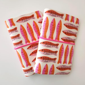 May include: Two fabric cases with a repeating pattern of orange and pink fish on a white background. The cases are bound with a bright pink elastic band. The fish are stylized with a simple design.