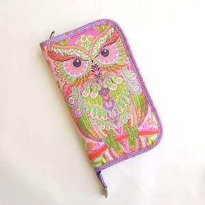 May include: A rectangular fabric wallet featuring a vibrant, multi-colored owl illustration with pink, green, orange, and purple tones. The wallet has a purple and white geometric patterned border and a metal zipper closure along the side.