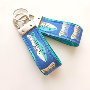 May include: Two fabric keychains featuring a blue background with a repeating pattern of light blue and white fish. Each keychain has a silver-toned metal clasp at the top and a teal green fabric backing, with visible stitching along the edges.