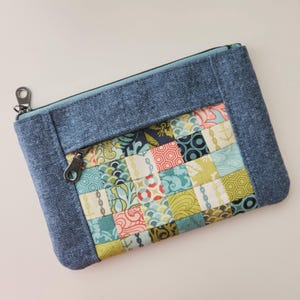 May include: A rectangular patchwork pouch with a denim blue border and zipper. The patchwork features squares of various patterns in teal, yellow, red, and navy blue. A silver-colored metal zipper pull and clasp are visible.