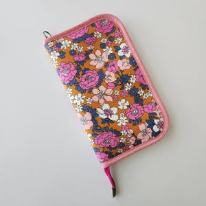 May include: A rectangular floral print case with a pink border and a pink pull tab. The case features a pattern of pink, white, and blue flowers on a golden-brown background. A small metal loop is attached to the top.