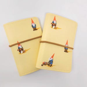 May include: Two yellow notebooks with brown elastic closures. Each notebook features a whimsical illustration of a garden gnome. The gnomes are depicted in various poses, including mowing a lawn and carrying tools. The notebooks are a charming stationery item.