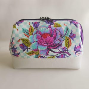May include: A zippered pouch featuring a light blue and white checkered background with a large, vibrant floral print in shades of purple, pink, and teal. The bottom section of the pouch is made of a solid white material with a smooth texture.