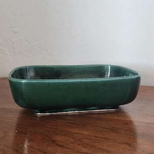 May include: A rectangular, dark green ceramic planter with rounded corners. The planter sits on a small, white-edged base. The glossy finish reflects light, highlighting its smooth surface. The planter is on a wooden surface.