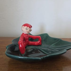 Puede incluir: Figura de cerámica vintage de un elfo sentado con un atuendo rojo, posado sobre un plato en forma de hoja verde oscuro. El elfo tiene una cara pintada con mejillas rosadas y un sombrero rojo. El plato es brillante.