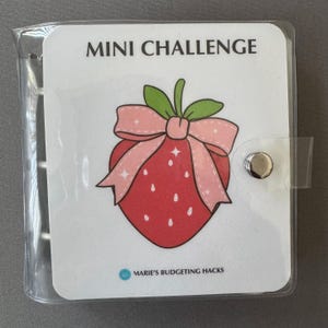 May include: A clear plastic mini binder with a white cover featuring a red strawberry illustration with a pink bow and the text "MINI CHALLENGE". The text "MARIE'S BUDGETING HACKS" is also visible.