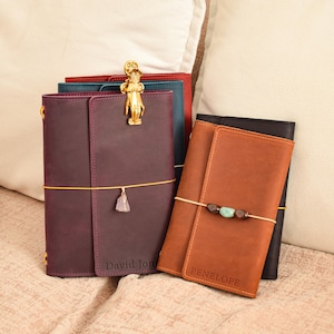 Puede incluir: Una colección de diarios de cuero en colores morado, azul, rojo, marrón y negro. El diario morado tiene un clip dorado con forma de mano y un pequeño dije de cristal. El diario marrón está personalizado con el nombre Penelope y el morado con David Jones.