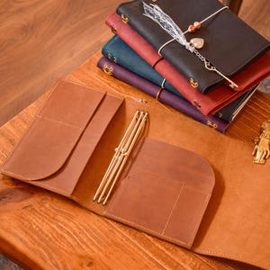Pode incluir: Uma capa de diário em couro castanho-claro com bolsos internos e elásticos para prender encartes. Uma pilha de quatro capas adicionais em preto, vermelho, azul e roxo, cada uma com um pingente de metal decorativo e fita de renda branca, está posicionada nas proximidades.