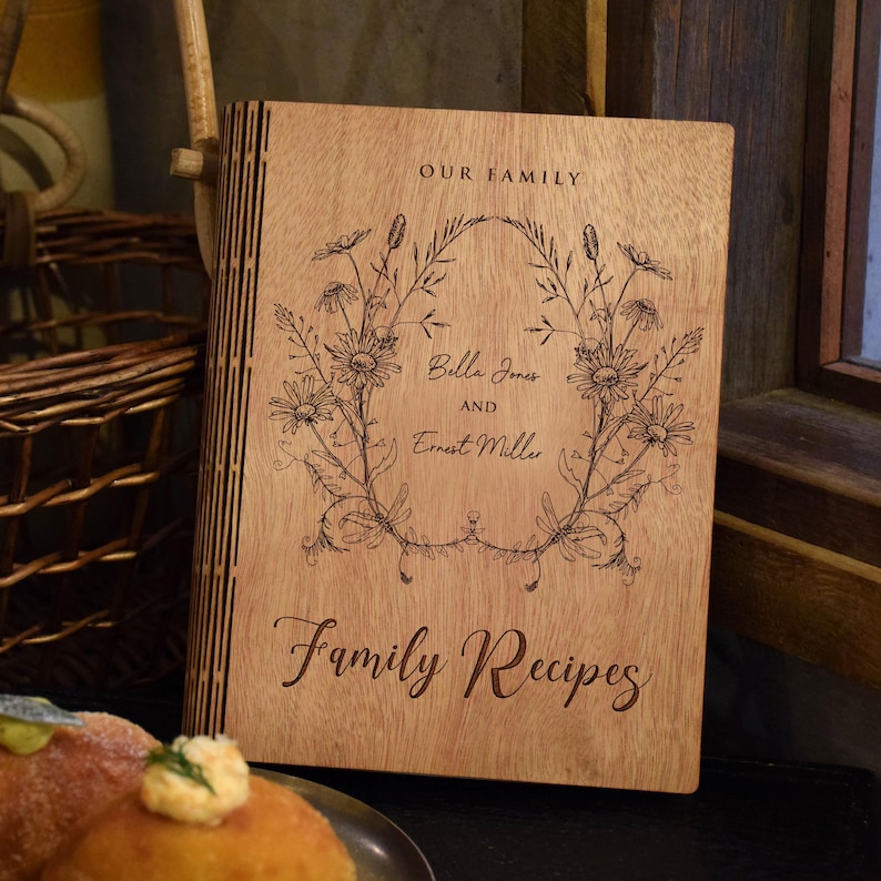 May include: A wooden family recipe book with an engraved floral design. The cover reads "OUR FAMILY" above a floral wreath with the names "Bella Jones and Ernest Miller" and "Family Recipes" below. The book is made of light brown wood.