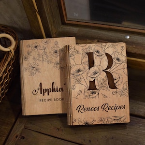 May include: Two wooden recipe books with floral designs and personalized text. One book says "Apphia Recipe Book" and the other says "Renees Recipes" with a large letter "R". The books are made of light-coloured wood.