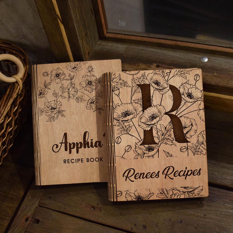 May include: Two wooden recipe books with floral designs and personalized text. One book is labeled "Apphia Recipe Book", the other features the letter "R" and the text "Renees Recipes". The books are made of light-coloured wood.