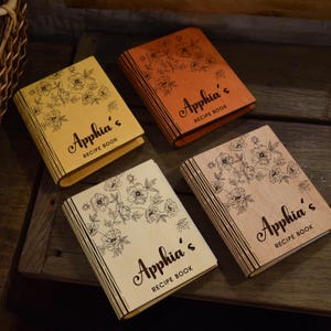 May include: Four wooden recipe books with floral designs and the name "Apphia's" on the cover. The books are in shades of yellow, orange, and natural wood tones. Each book has a unique wood grain.