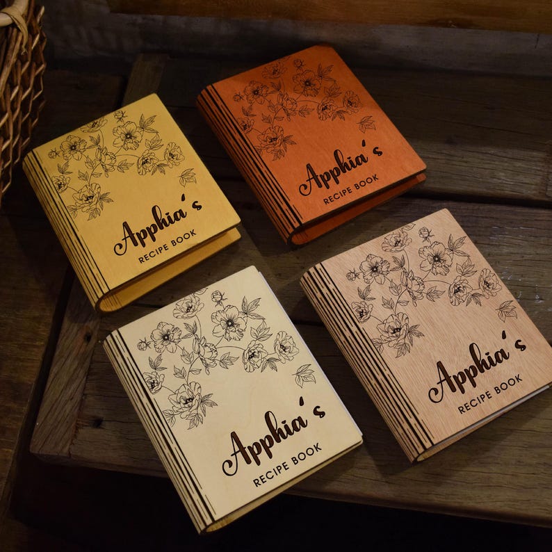 May include: Four wooden recipe books with floral designs and the name "Apphia's" on the cover. The books are in shades of yellow, orange, and natural wood tones. Each book has a unique wood grain.
