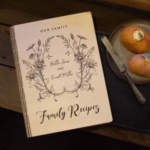 May include: A wooden recipe book with the words "OUR FAMILY" at the top and "Family Recipes" at the bottom. The cover features a floral wreath design with the names "Bella Jones and Ernest Miller" in the center. A plate with two pastries and a knife is on the right.