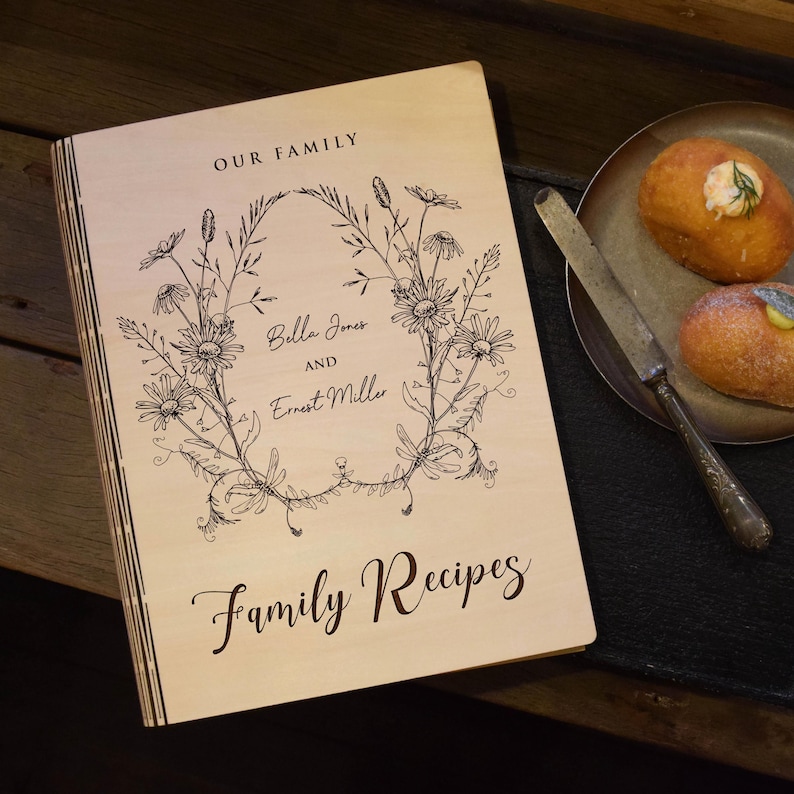 May include: A wooden recipe book with the words "OUR FAMILY" at the top and "Family Recipes" at the bottom. The cover features a floral wreath design with the names "Bella Jones and Ernest Miller" in the center. A plate with two pastries and a knife is on the right.