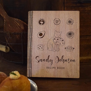 May include: Wooden recipe book with engraved food illustrations and a cat holding a donut. The cover displays the name "Sandy Johnson" and "Recipe Book" in a script font. The book is made of light-coloured wood.