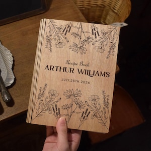 Könnte beinhalten: Ein hölzernes Rezeptbuch mit floralen Gravuren und dem Text "Recipe Book ARTHUR WILLIAMS JULY.28TH.2026". Das Buch ist geöffnet und zeigt das eingravierte Design auf dem Einband. Das Buch ist aus hellem Holz.