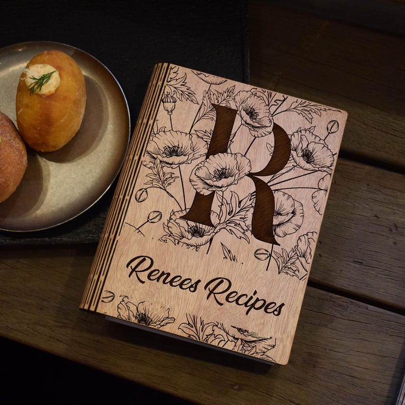 May include: A wooden recipe book featuring a poppy floral design and a dark brown letter "R". The cover is inscribed with "Renees Recipes" in a cursive script. The book rests on a wooden surface, with food on a plate visible in the background.