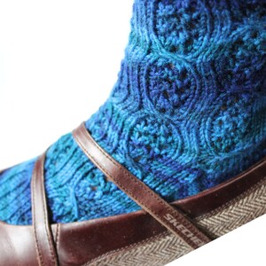 May include: Blue and teal knitted socks with a swirling pattern, worn with brown leather shoes with a brown and white herringbone pattern. The shoes have the word "SKECHERS" on the side.