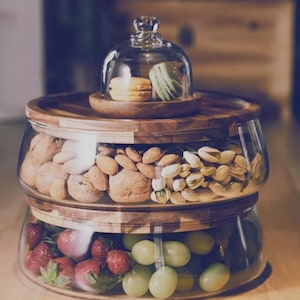 Peut inclure: Un plat de service à plusieurs niveaux avec un dôme en verre. Le niveau supérieur contient deux macarons. Le niveau intermédiaire est rempli d'amandes, de noix et de pistaches. Le niveau inférieur contient des fraises et des raisins verts. Le plat a des accents en bois.