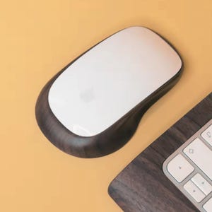 Puede incluir: Un ratón de ordenador blanco y de madera oscura con el logotipo de Apple. El ratón tiene un diseño curvo y está sobre un fondo amarillo. Una parte de un teclado es visible en la esquina inferior derecha.