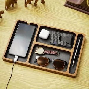 Peut inclure: Un plateau organisateur en bois avec des compartiments pour un téléphone, une montre, des lunettes de soleil, un portefeuille et un stylo. Le plateau est en bois brun clair et est doublé de feutre noir.