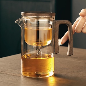 Może przedstawiać: Szklany czajnik do herbaty z drewnianym uchwytem i pokrywką. Czajnik ma wbudowany filtr do herbaty i jest wypełniony herbatą.