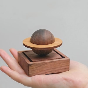 Op de afbeelding: Een houten bol met een donkerbruine afwerking zit op een kleiner houten platform. De bol wordt omringd door een dunne, lichtbruine ring.