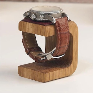 Op de afbeelding: Een houten horloge standaard met een bruin leren horlogeband. De standaard is gemaakt van lichtbruin hout en heeft een gebogen ontwerp.
