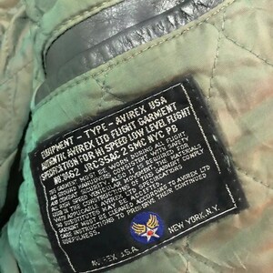 Może przedstawiać: Czarna sk&oacute;rzana metka z białym tekstem, kt&oacute;ry brzmi "Equipment - Type - Avirex USA Authentic Avirex Ltd Flight Garment Specification for Hi Speed Low Level Flight No: 3462 JRC35AC 2 SMC NYC PB". Metka zawiera r&oacute;wnież niebiesko-białą naszywkę z gwiazdą i skrzydłami.