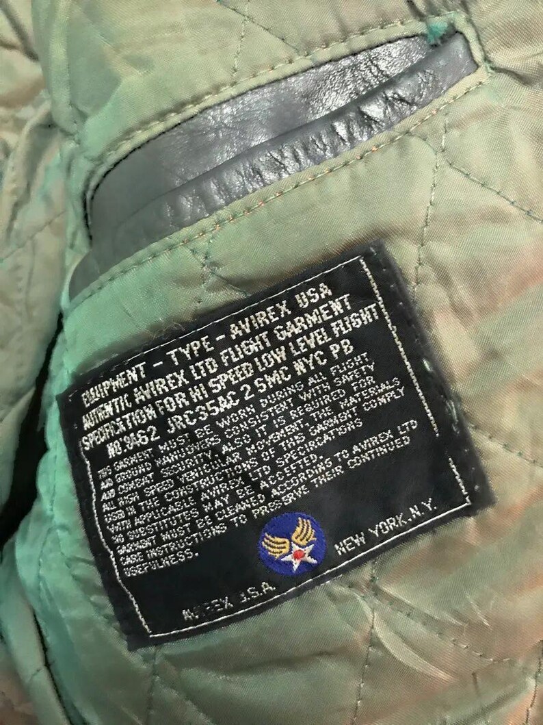 Może przedstawiać: Czarna sk&oacute;rzana metka z białym tekstem, kt&oacute;ry brzmi "Equipment - Type - Avirex USA Authentic Avirex Ltd Flight Garment Specification for Hi Speed Low Level Flight No: 3462 JRC35AC 2 SMC NYC PB". Metka zawiera r&oacute;wnież niebiesko-białą naszywkę z gwiazdą i skrzydłami.