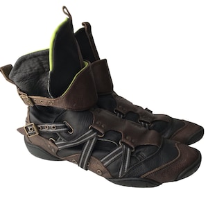 May include: A pair of high-top leather boots in black and brown. The boots feature multiple straps with buckles, a folded-over collar, and a gray and white striped accent. The interior lining is black with a neon yellow trim.