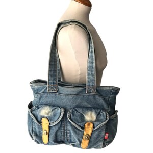 May include: A blue denim tote bag with two front pockets, each secured by a tan leather strap and button closure. The bag has two denim handles and a red tag on the side. The bag is displayed on a mannequin.