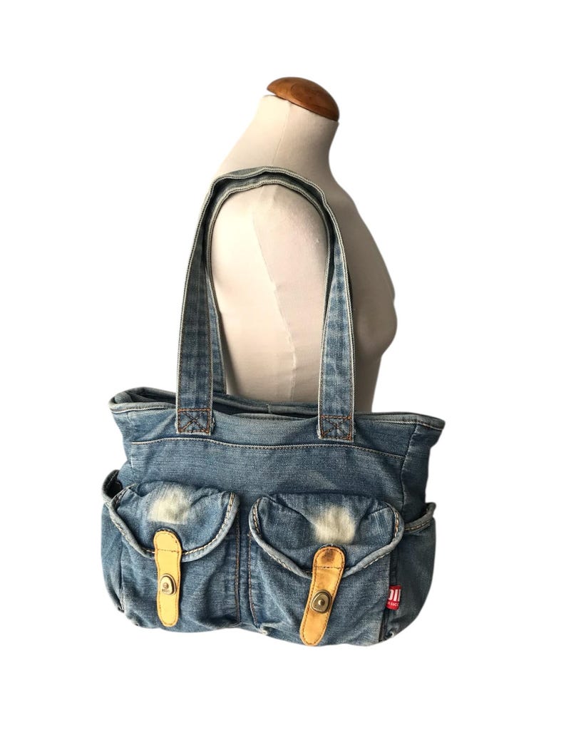 May include: A blue denim tote bag with two front pockets, each secured by a tan leather strap and button closure. The bag has two denim handles and a red tag on the side. The bag is displayed on a mannequin.