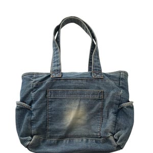 May include: A blue denim tote bag with two handles and a front pocket. The bag has a faded appearance and side pockets. The denim material is a medium wash, and the bag appears to be new.