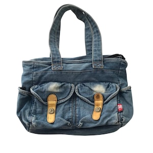 May include: A denim tote bag with two front pockets and tan leather-like closures. The bag has two denim handles and a small red and white tag on the side. The bag is made of faded blue denim.