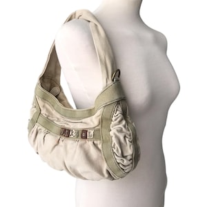 May include: A beige hobo-style shoulder bag made of canvas fabric with gathered side panels and olive green trim. The front features a horizontal strap with the word DIESEL spelled out in individual brown and white fabric letter patches.