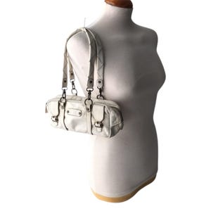 K&ouml;nnte beinhalten: Eine cremefarbene Schultertasche aus Kunstleder mit rechteckiger Form. Die Tasche hat zwei kurze Griffe und einen langen, verstellbaren Riemen. Sie verf&uuml;gt &uuml;ber mehrere Taschen mit Schnallenverschl&uuml;ssen und Metallbeschl&auml;gen. Die Tasche wird auf einer Schaufensterpuppe pr&auml;sentiert.