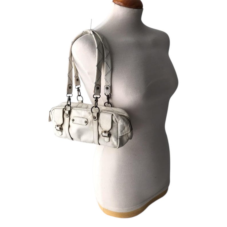 K&ouml;nnte beinhalten: Eine cremefarbene Schultertasche aus Kunstleder mit rechteckiger Form. Die Tasche hat zwei kurze Griffe und einen langen, verstellbaren Riemen. Sie verf&uuml;gt &uuml;ber mehrere Taschen mit Schnallenverschl&uuml;ssen und Metallbeschl&auml;gen. Die Tasche wird auf einer Schaufensterpuppe pr&auml;sentiert.