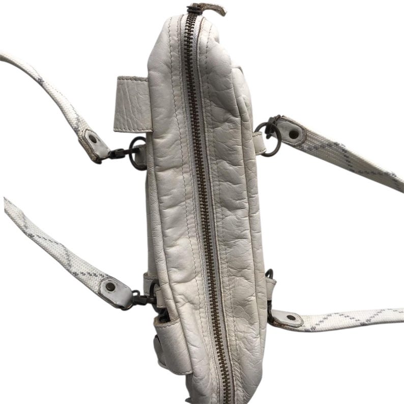 K&ouml;nnte beinhalten: Cremefarbene Messenger-Tasche aus Leder mit Rei&szlig;verschluss und mehreren F&auml;chern. Die Tasche hat eine strukturierte Oberfl&auml;che und einen langen, verstellbaren Riemen mit geometrischem Muster. Metallbeschl&auml;ge akzentuieren das Design der Tasche.