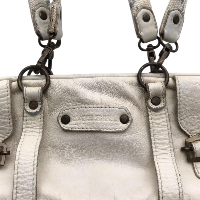K&ouml;nnte beinhalten: Cremefarbene Lederhandtasche mit dem Markennamen "DIESEL" auf der Vorderseite. Die Tasche hat zwei obere Griffe mit gemusterten Riemen und Metallbeschl&auml;gen. Die Tasche hat eine rechteckige Form und ein leicht abgenutztes Aussehen.