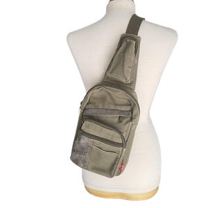 May include: An olive green canvas sling bag with multiple pockets and a shoulder strap. The bag features the brand name "DIESEL" printed on the front and a small red tag. The bag is displayed on a mannequin.