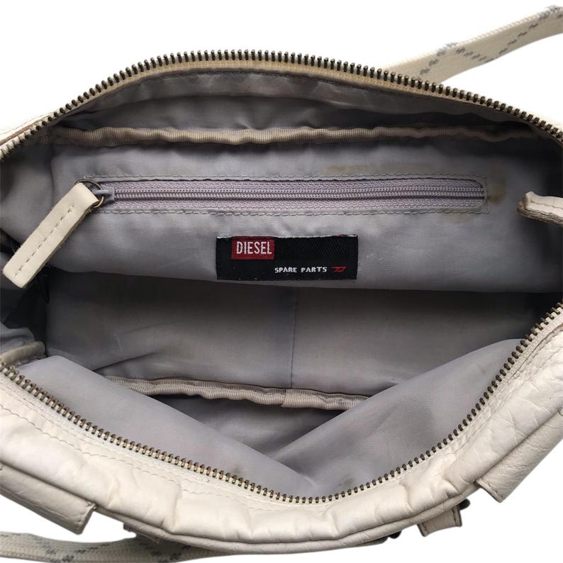 K&ouml;nnte beinhalten: Cremefarbene Diesel-Handtasche mit Rei&szlig;verschluss und grauem Innenraum. Die Tasche hat eine kleine Innentasche mit Rei&szlig;verschluss und ein Diesel-Etikett mit der Aufschrift "SPARE PARTS". Die Tasche hat eine strukturierte Au&szlig;enseite und einen Schulterriemen.