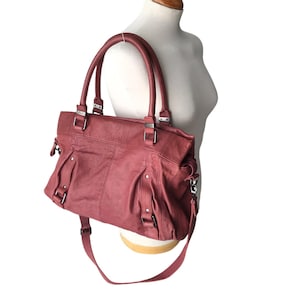 May include: A dusty rose handbag featuring two top handles and a detachable shoulder strap. The bag has a textured surface with vertical stitched panels, silver-toned metal hardware, and side buckles. It is displayed on a white mannequin torso.