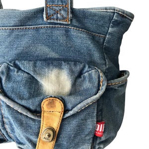 May include: A denim tote bag with a light blue wash. The bag features a front pocket with a tan leather strap and a metal button closure. The word "DIESEL" is visible on the strap. A red tag with white text is attached to the side.