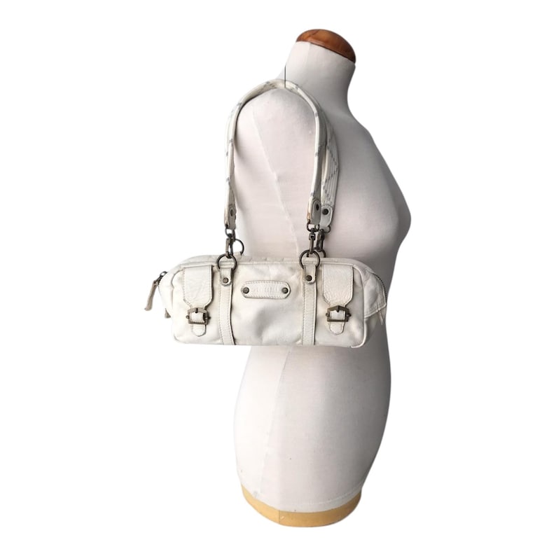 K&ouml;nnte beinhalten: Eine wei&szlig;e, rechteckige Handtasche mit oberem Griff und Schulterriemen wird auf einer Schaufensterpuppe pr&auml;sentiert. Die Tasche hat zwei kleine Schnallen, einen Rei&szlig;verschluss und Metallbeschl&auml;ge. Die Tasche ist ca. 30 cm lang und 15 cm hoch.