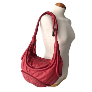 May include: A red shoulder bag with a curved shape and a knotted strap. The bag has a zipper closure and a dark metal ring detail. The bag is displayed on a mannequin.