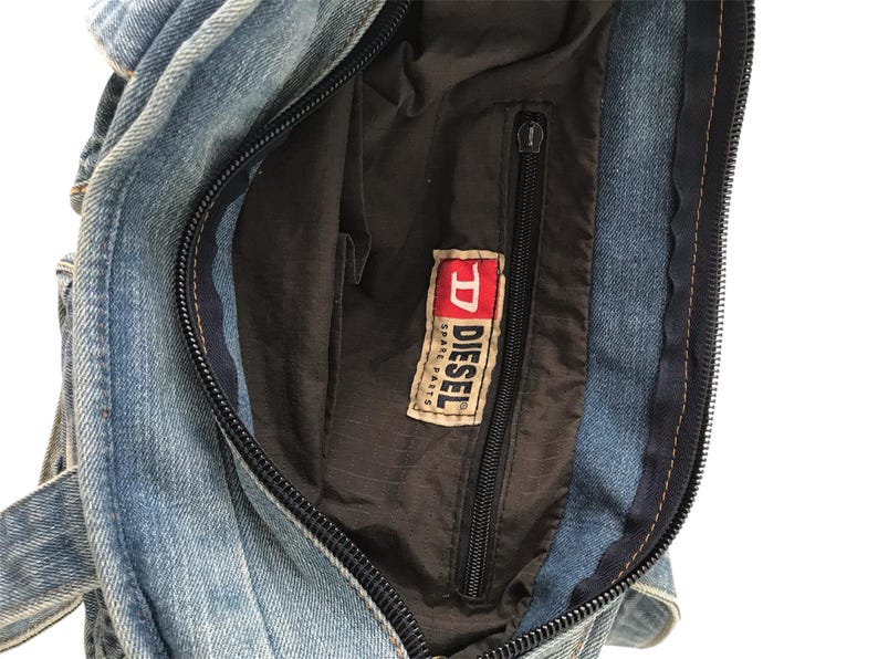 May include: A denim handbag with a black zipper and a black interior. The bag features a Diesel Spare Parts label. The denim is a light blue wash.