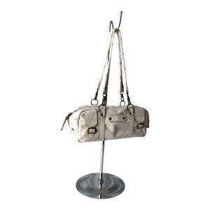K&ouml;nnte beinhalten: Cremefarbene Lederhandtasche mit rechteckiger Form, zwei Henkeln und Rei&szlig;verschluss. Die Tasche hat dekorative Schnallen und h&auml;ngt an einem silbernen Pr&auml;sentationsst&auml;nder. Die Tasche ist neutralfarben und scheint neu zu sein.