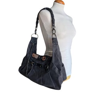 May include: A dark gray hobo bag featuring a braided shoulder strap, silver-tone metal hardware, and a decorative chain accent. The bag includes multiple metal rings, studs, and rectangular metal plates along the top edge with a slouchy silhouette.